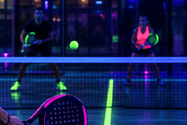 Deux joueurs de padel prêts derrière le filet, balle vert fluo en vol et ligne fluorescente sur la piste